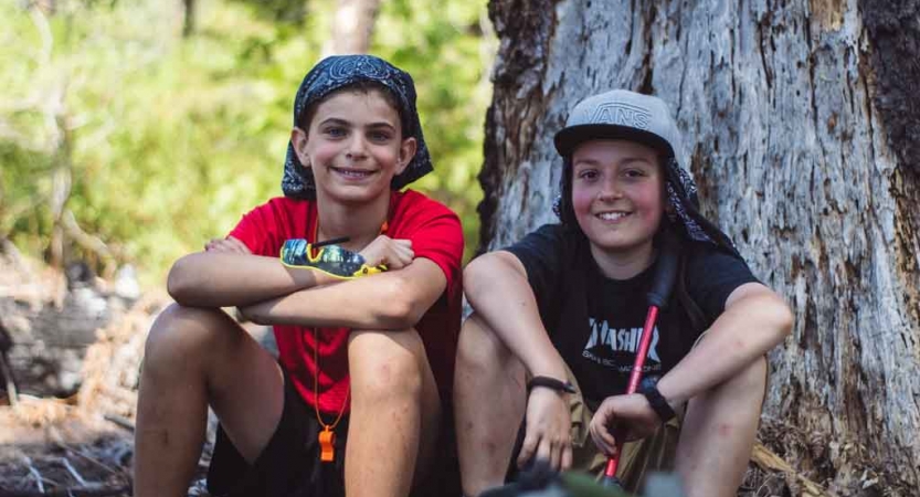 Two young people sit beside the trunk of a tree and smile for the photo 